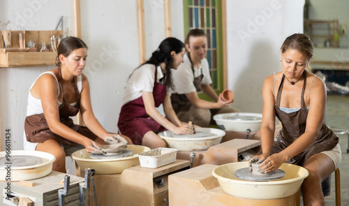 Positive young lady wearing apron modeling clay dish from row material using pottery wheel in taller workroom