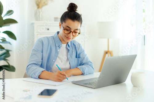 Focused successful arabian or indian brunette woman in a blue shirt, sitting at a work desk while working in the laptop, doing online tasks, develop new digital project, planning marketing strategy