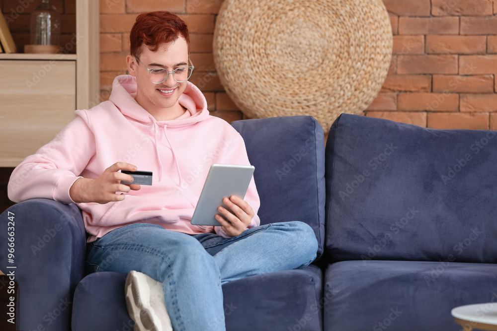 Obraz premium Handsome young man with credit card and tablet computer sitting on sofa while shopping online
