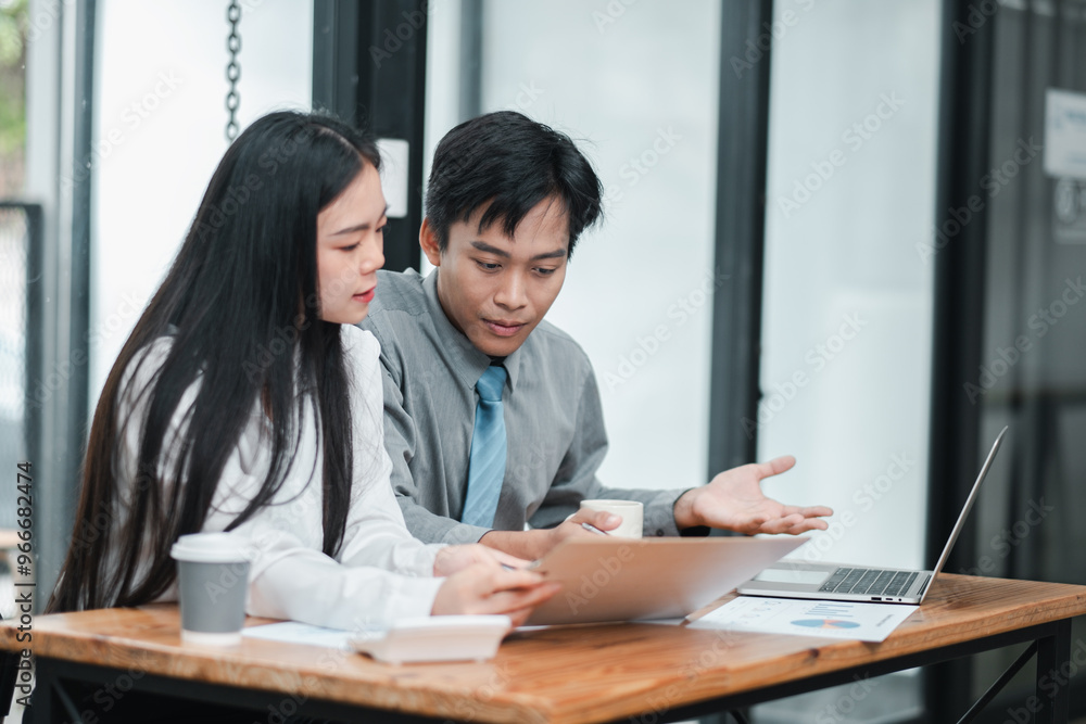 Two young professionals working together in a modern office, discussing a project with laptops and documents on a wooden table. Two young professionals working together in a modern office, discussing a project with laptops and documents on a wooden table.