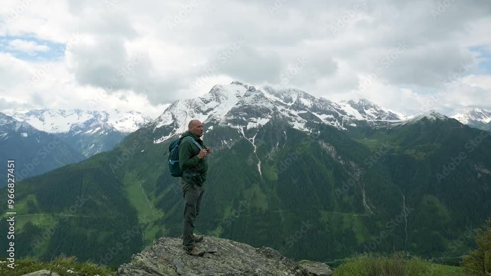 A man with a backpack stands on a mountain peak, overlooking a stunning landscape of snow-covered mountains and green valleys. A scene capturing the beauty of nature and adventure. 4k footage A man with a backpack stands on a mountain peak, overlooking a stunning landscape of snow-covered mountains and green valleys. A scene capturing the beauty of nature and adventure. 4k footage