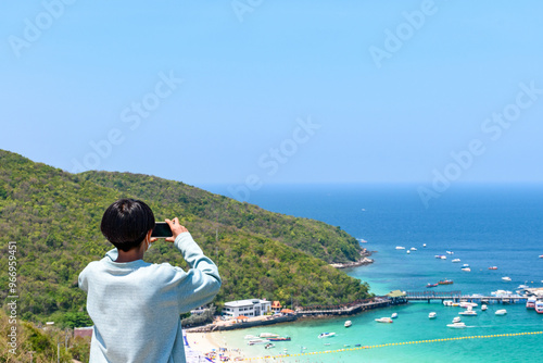 Man holding a smartphone is taking a photo of the view of a tropical beach and an island in the sea on a mountaintop. White sandy beach view Turquoise water at Koh Larn Islands, Thailand.