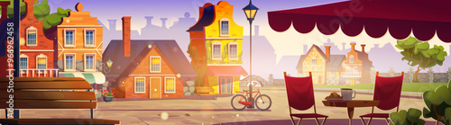 Old Europe street with cafe table with coffee cup and croissant, cute houses and park with stone bridge and fence, green trees and bicycle near streetlight, wooden bench under morning sun light.