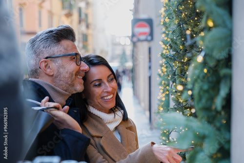 Happy couple enjoys the festive season shopping in the city streets filled with christmas lights and decorations, laughing and looking at holiday displays while carrying bags