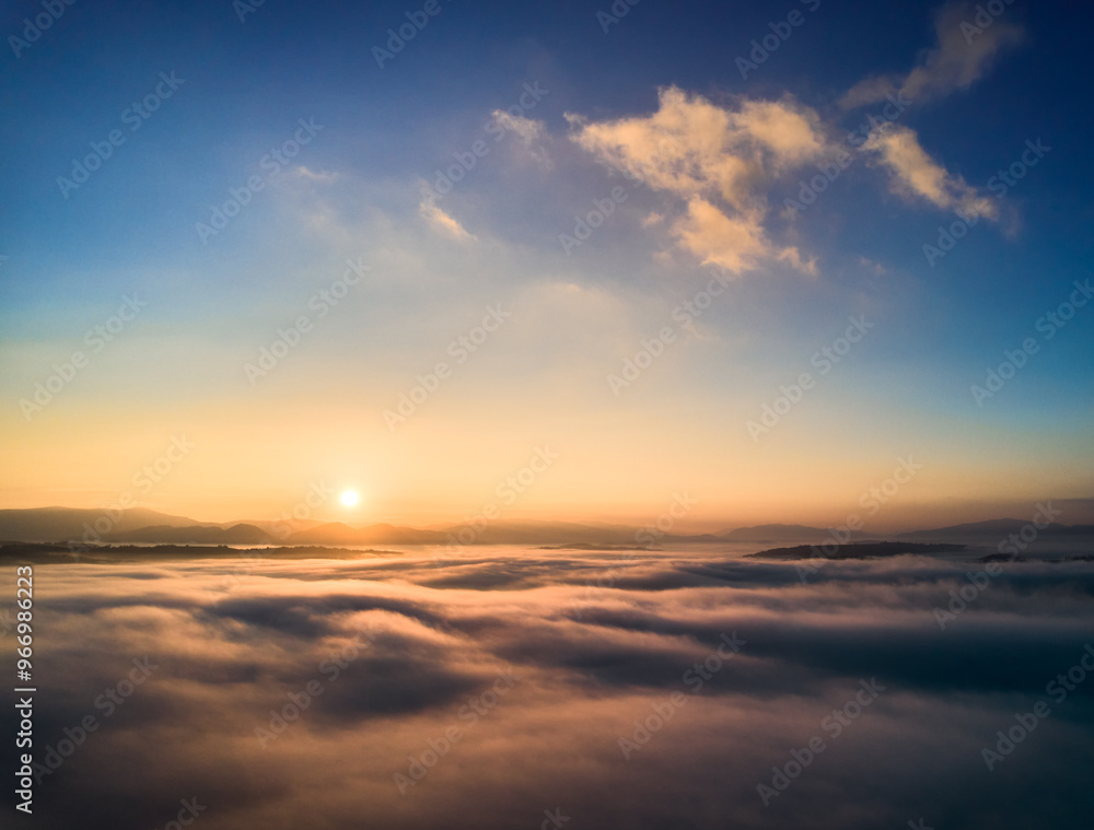 Aerial view of sun rises above sea of clouds, painting sky with soft hues of orange and blue. Light, wispy clouds scatter across horizon, capturing serene and beauty of dawn breaking over landscape. Aerial view of sun rises above sea of clouds, painting sky with soft hues of orange and blue. Light, wispy clouds scatter across horizon, capturing serene and beauty of dawn breaking over landscape.