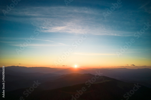 Sun sets behind rolling hills, casting warm, golden glow across sky. Wispy clouds, tinged with hues of orange and yellow, stretch across deepening blue sky. Serene and expansive landscape.