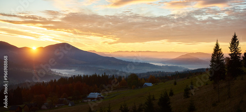 Sun rises over rolling hills, casting warm golden glow across landscape. Soft mist blankets valley, colors decorate trees. Silhouetted pine trees and few scattered homes add depth morning scene.