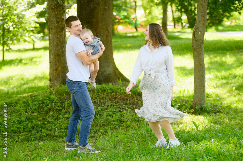 Fototapeta premium In public park, man and woman lovingly hold tiny baby in their arms, surrounded by bright green trees and grass, having fun, dancing, spinning. Summer fun for families in nature