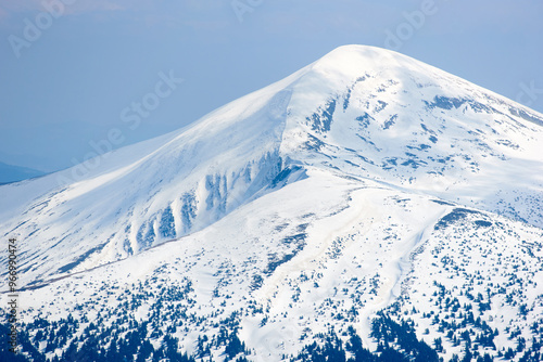 Snow-covered mountain peak under clear blue sky, with patches of snow on slopes and scattered trees. Serene winter landscape contrasts sharply with the dark, forested areas below. Hoverla, Ukraine photo