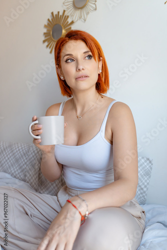 Red-haired woman sits on a bed, holding a coffee cup and gazing thoughtfully into the distance. She is dressed in a white tank top and beige pants