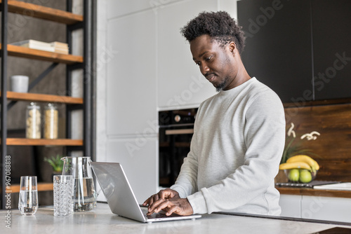 African American man stands at kitchen, working remotely on laptop photo