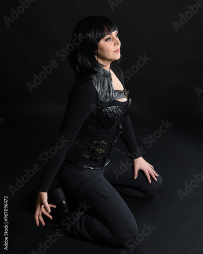 Full length portrait of beautiful black female model wearing black sci fi futuristic dystopian leather bodysuit costume. Kneeling pose. isolated dark studio background moody silhouette.