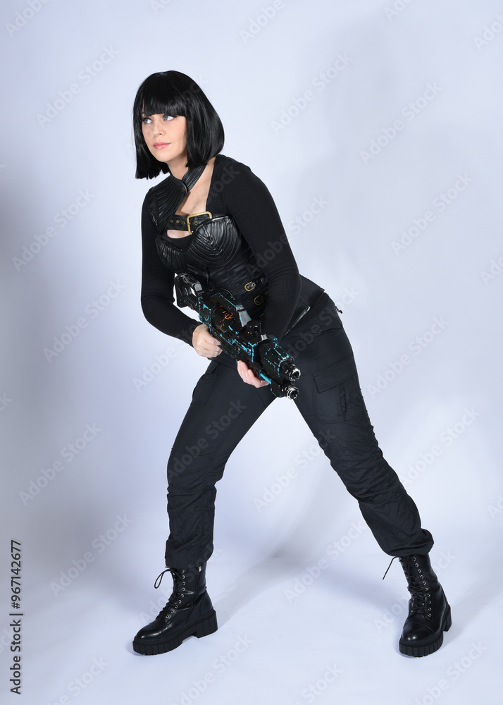 Naklejka premium Full length portrait of beautiful black female model with short black hair wearing sci fi futuristic dystopian leather bodysuit costume. Standing pose holding gun weapon, isolated white studio backgro