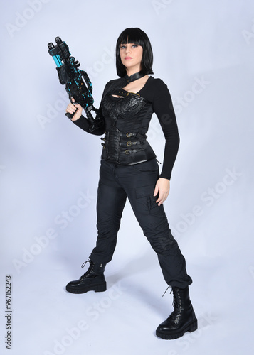 Full length portrait of beautiful black female model with short black hair wearing sci fi futuristic dystopian leather bodysuit costume. Standing pose holding gun weapon, isolated white studio backgro