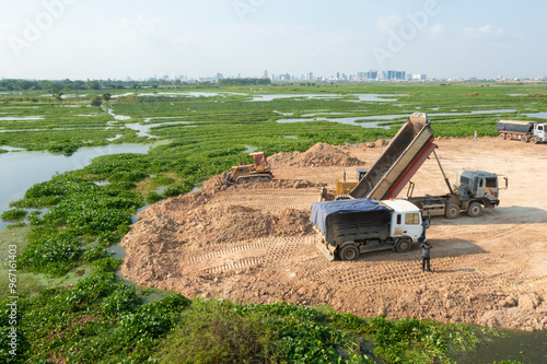 Construction Site with Dump Trucks Unloading Soil Near Green Wetlands
