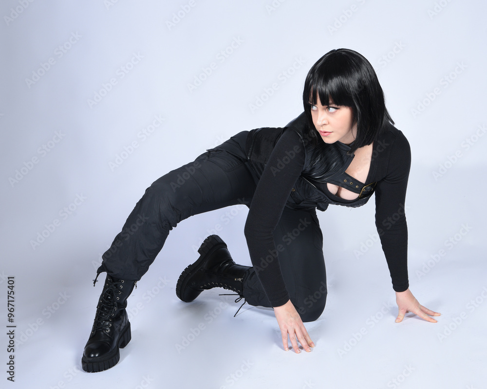 Full length portrait of beautiful black female model with short black hair wearing sci fi futuristic dystopian leather bodysuit costume. Relaxed sitting pose, isolated white studio background. Full length portrait of beautiful black female model with short black hair wearing sci fi futuristic dystopian leather bodysuit costume. Relaxed sitting pose, isolated white studio background.