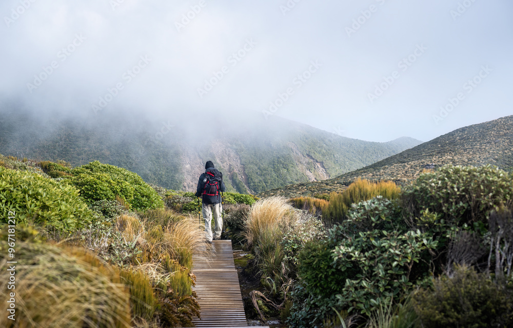 Man hiking Pouakai circuit, walking on boardwalk. Clouds drifting over the mountains. Egmont National park. New Zealand. Man hiking Pouakai circuit, walking on boardwalk. Clouds drifting over the mountains. Egmont National park. New Zealand.