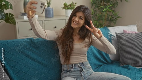 A beautiful young woman takes a selfie in her living room, flashing a peace sign and smiling while sitting on a sofa, surrounded by plants and modern decor. photo