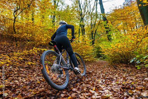 Woman wearing black sporty jacket, black cycling pants and white bike helmet riding her bike uphill in forest in autumn scenery. Back view.