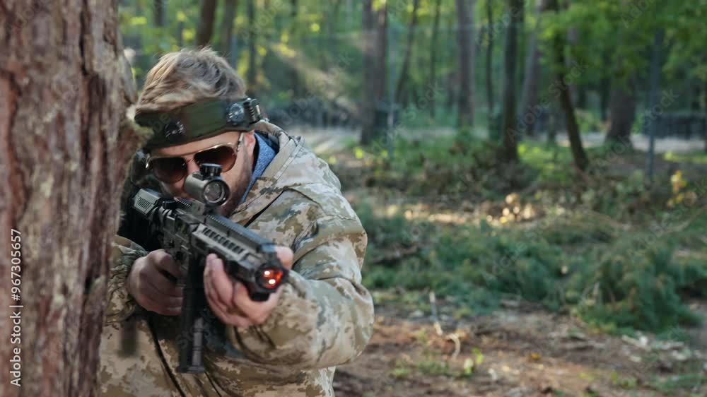 Shoots enemy, tree stance, enemy battle. Man adorned in camouflage gear with marker on his head leans against tree, accurately aiming Laser tag gun and hitting opponent in forest setting. Shoots enemy, tree stance, enemy battle. Man adorned in camouflage gear with marker on his head leans against tree, accurately aiming Laser tag gun and hitting opponent in forest setting.