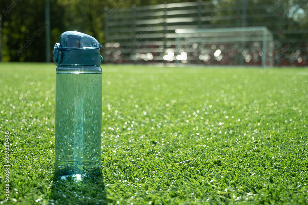 Transparent refreshing blue bottle of water in sunny day with shadow on outdoor soccer sports ground with artificial grass covering. Concept water balance for workout. Copy space. Transparent refreshing blue bottle of water in sunny day with shadow on outdoor soccer sports ground with artificial grass covering. Concept water balance for workout. Copy space.