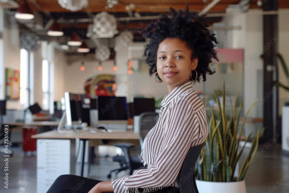 A woman with natural hair and hoop earrings, seated in a contemporary office space, smiles confidently towards the camera. A woman with natural hair and hoop earrings, seated in a contemporary office space, smiles confidently towards the camera.