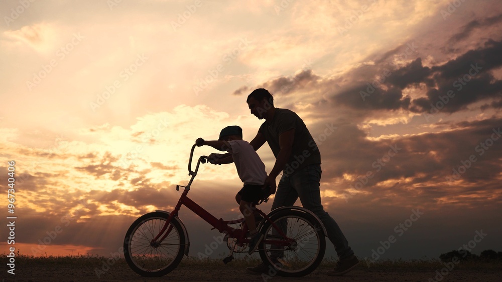 Fototapeta premium happy family in the park. father teaching son to ride a bike at sunset silhouette in the park. son child learning to ride a bike at sunset father helping son. child playing riding lifestyle a bike