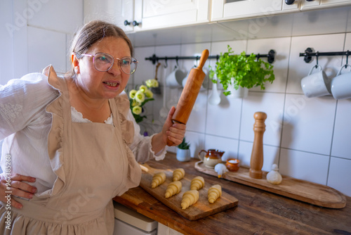 woman housewife, housekeeper, angry wife in linen old-fashioned apron holding wooden rolling pin, threatening household, stress from household routine, kitchen interior photo