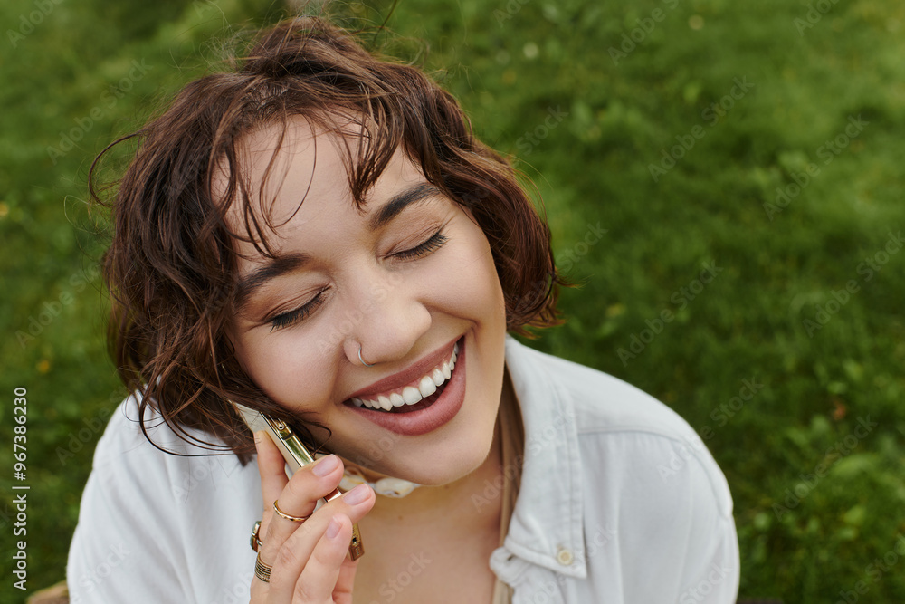 A cheerful young woman enjoys a summer picnic, laughing while chatting on her phone amidst green grass. A cheerful young woman enjoys a summer picnic, laughing while chatting on her phone amidst green grass.