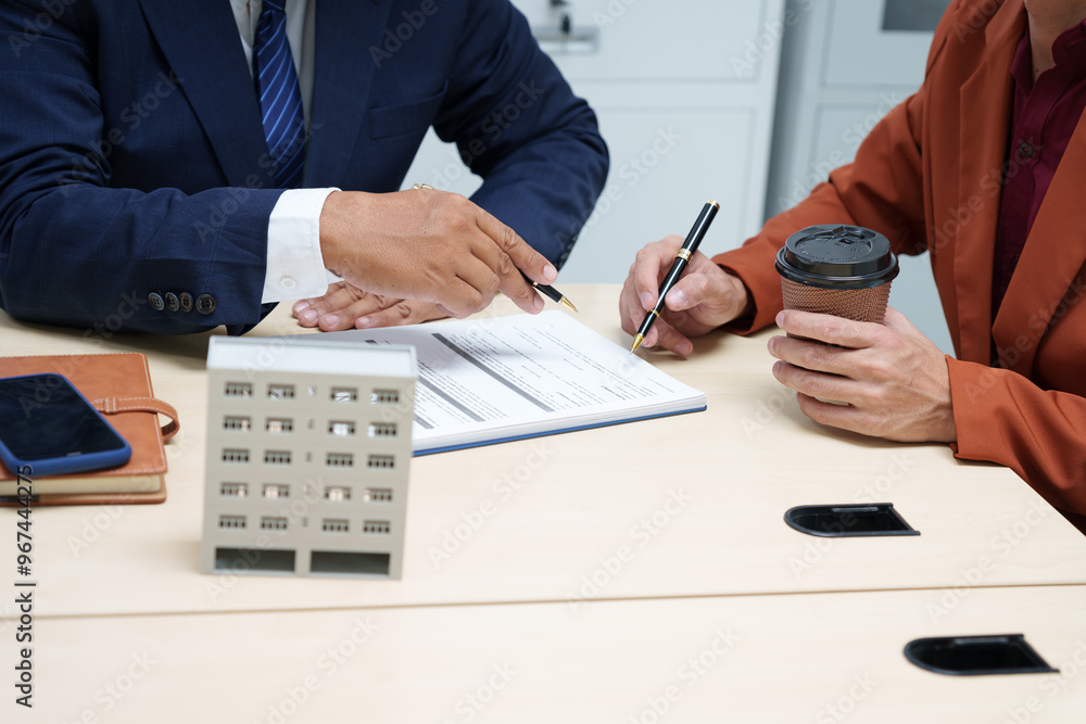 Fototapeta premium A businessman at a desk in a sales office sells a condo building to a customer. They agree to sign a contract, covering real estate investments in offices, apartments, and commercial properties.