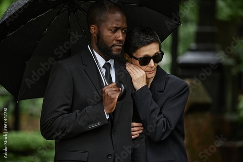 Medium shot of mourning multiethnic couple grieving at memorial ceremony. Senior woman clinging to Black mans shoulder hiding from rain under black umbrella at cemetery