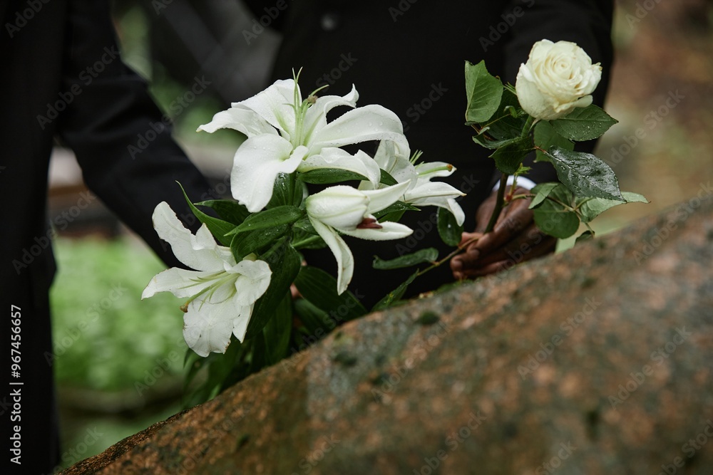 © Seventyfour - Close up on rain wet white lilies and rose in hands of two unrecognizable people placing flowers on stone memorial monument to commemorate deceased loved one at cemetery, copy space © Seventyfour - Close up on rain wet white lilies and rose in hands of two unrecognizable people placing flowers on stone memorial monument to commemorate deceased loved one at cemetery, copy space