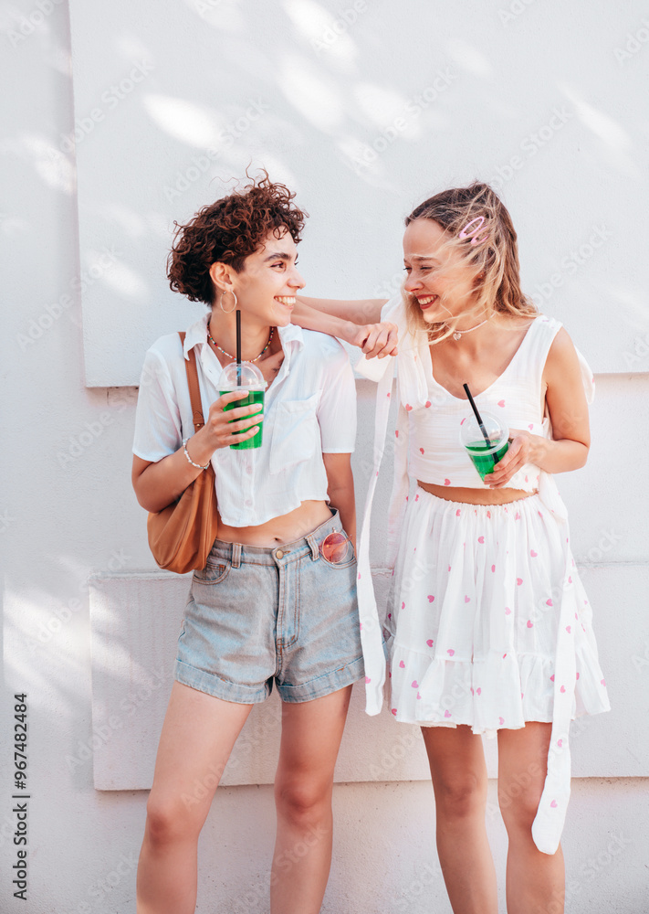 Two young beautiful smiling hipster female in trendy summer clothes. Carefree women posing outdoors. Positive models holding and drinking fresh cocktail smoothie drink in plastic cup Two young beautiful smiling hipster female in trendy summer clothes. Carefree women posing outdoors. Positive models holding and drinking fresh cocktail smoothie drink in plastic cup