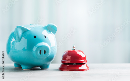 A close-up of a red service bell next to a piggy bank on a wooden table against a bright white background. The image represents a call to action for financial planning and savings. photo