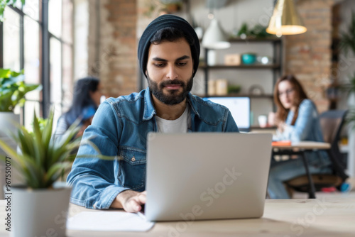 Portrait of handsome Arab businessman wearing hat and casual clothes while working in open space, modern workplace or cafe, islamic person. Bearded muslim man working on laptop in office