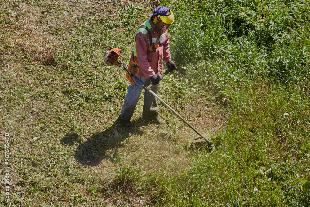A gardener in coveralls and protective overalls cuts overgrown grass with a lawnmower. A gardener in coveralls and protective overalls cuts overgrown grass with a lawnmower.