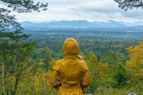 The scenic beauty of the valley and mountains is admired by a woman wearing a yellow raincoat on a hill.