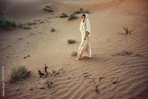 Woman in flowing white garment amid desert landscape. Sparse vegetation adds texture. Her presence exudes mystery and grace. Ideal for themes of solitude, nature connection, and cultural exploration. photo