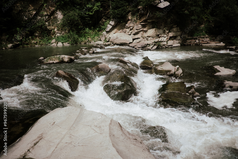 Fototapeta premium A serene river flowing over rocks in a lush green landscape with scattered boulders on a sunny day