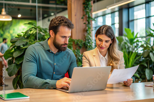 Two Colleagues Collaborating in a Modern Office, Reviewing Documents on a Laptop with Greenery Backdrop