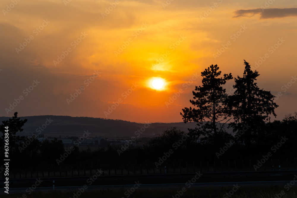 Orange sunset sun getting down amongs the trees over the road in the shade. Dramatic orange sky during the susnset.
