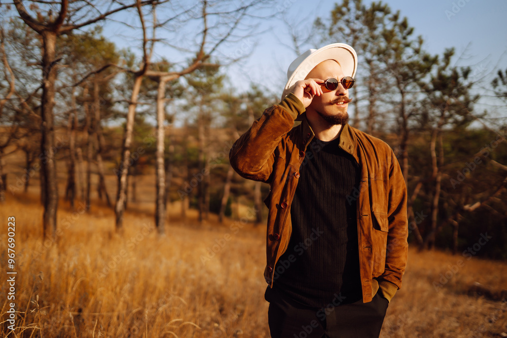 Man wearing sunglasses and a large hat enjoys a sunny day outdoors in a grassy area surrounded by trees during autumn. Autumn scenery. Man wearing sunglasses and a large hat enjoys a sunny day outdoors in a grassy area surrounded by trees during autumn. Autumn scenery.