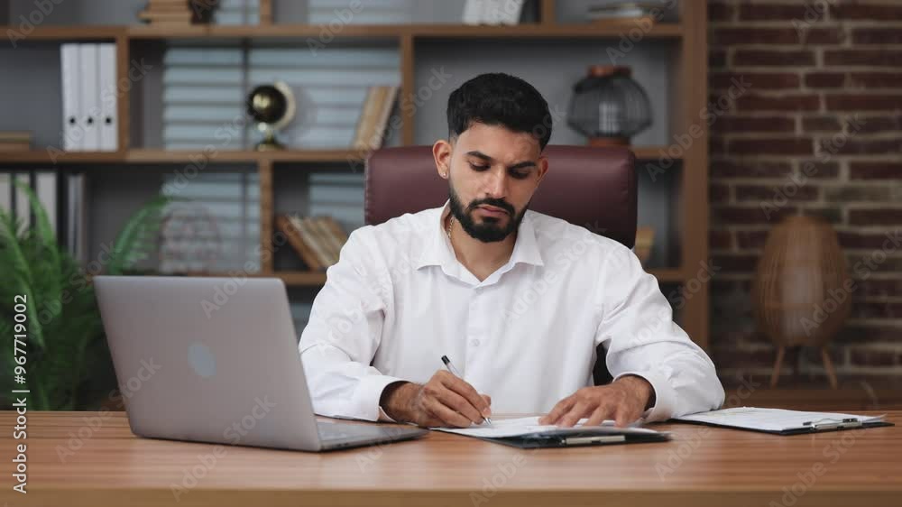 Front view of hardworking indian male typing on keyboard of modern laptop in office corpotate. Serious man dressed in white shirt making notes on documents indoors. Front view of hardworking indian male typing on keyboard of modern laptop in office corpotate. Serious man dressed in white shirt making notes on documents indoors.
