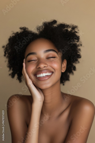 Joyful woman, her hand resting on her cheekbone, bare shoulders glowing against a neutral beige background, smiling brightly.