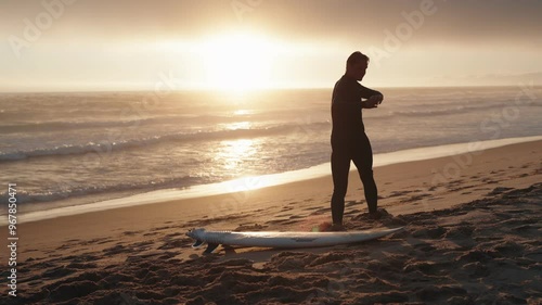 A surfer stretches his arms on the beach with his board at sunset, preparing to enter the water.