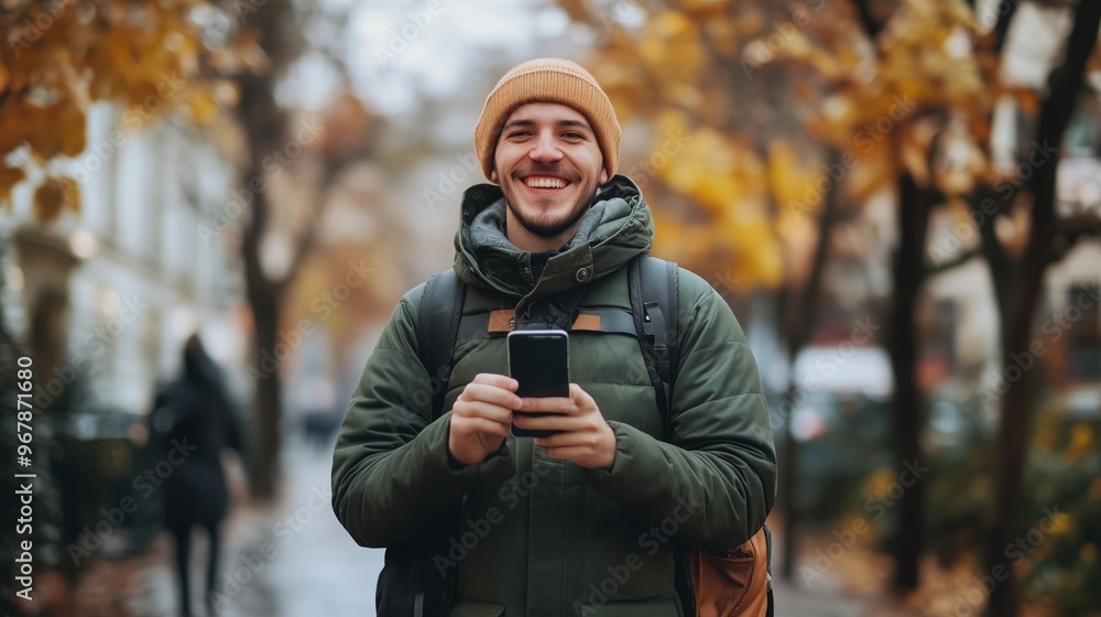 Young man smiling while checking his smartphone during autumn in a city street lined with colorful trees Young man smiling while checking his smartphone during autumn in a city street lined with colorful trees
