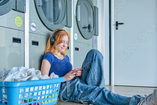 Redhead girl in a laundry looking at her smart phone while waiting for the clothes to be washed