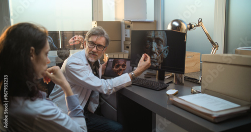 Two diverse scientists discuss 3D model of human head anatomy displayed on PC and tablet computer screens, conduct scientific research. Archaeologists or paleontologists work in archaeological lab. photo