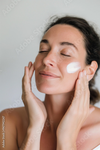 Woman applying facial moisturizer lotion on clean skin, gray background 