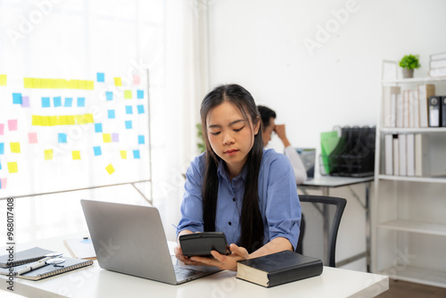 Financial Focus Amidst Office Hustle: A young Asian professional intently calculates expenses, her brow furrowed in concentration, highlighting the demands of modern business. 